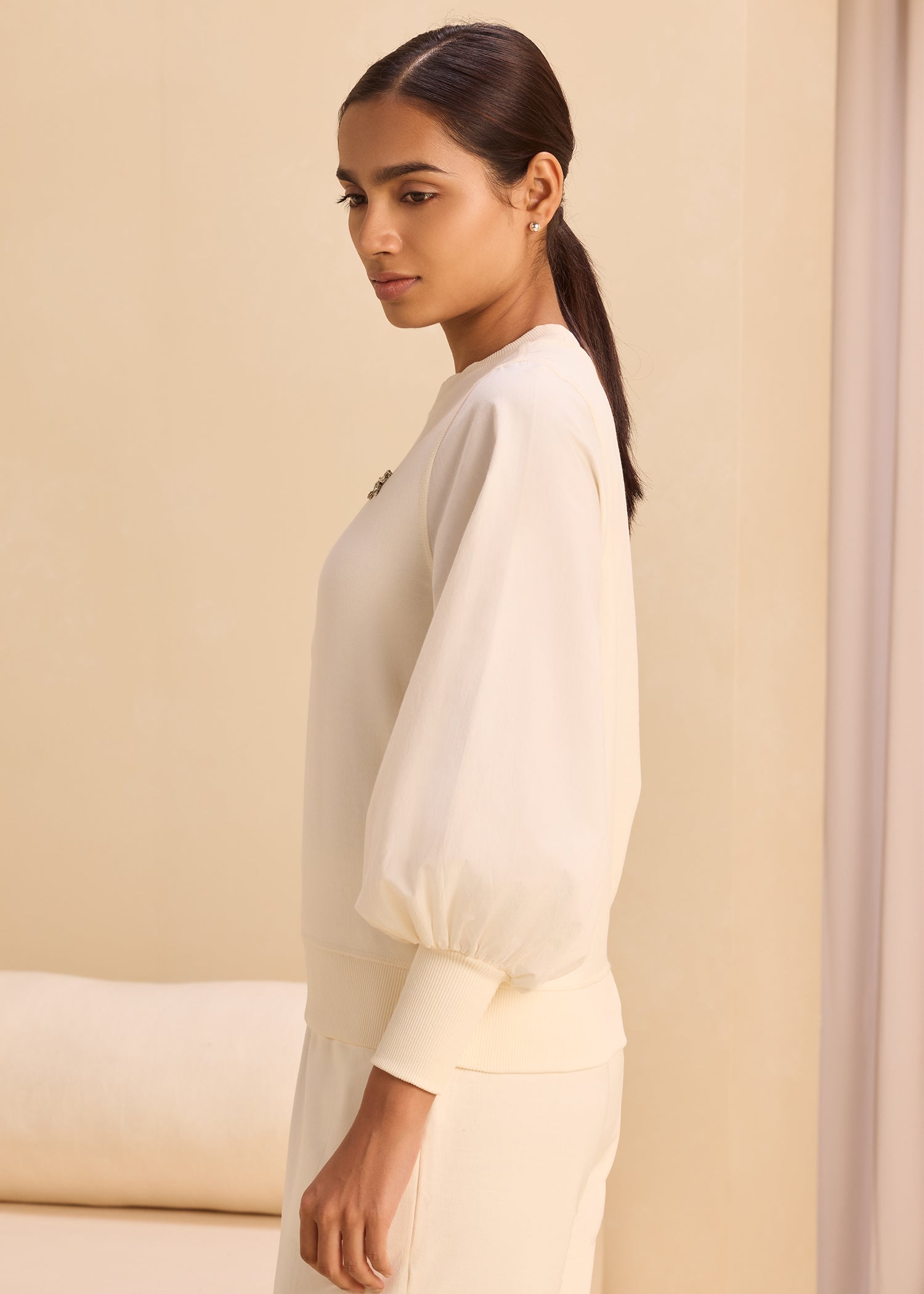 Young woman in cream outfit standing sideways indoors with thoughtful expression