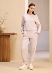 Woman in a light beige lounge set, including a long-sleeve sweatshirt and joggers, standing indoors next to a wooden table with a ceramic vase.