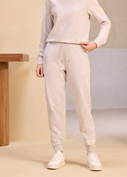 Full-length image of a person wearing a grey lounge set with a drawstring hoodie and joggers, paired with white chunky sneakers, standing in a soft, neutral-colored minimalist setting.
