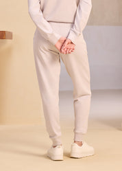Back view of a person wearing light beige joggers and white sneakers standing on a minimalist beige background.