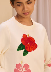 Close-up of a model wearing a cream sweatshirt featuring a large red embroidered hibiscus flower with green leaves, displaying intricate detailing.